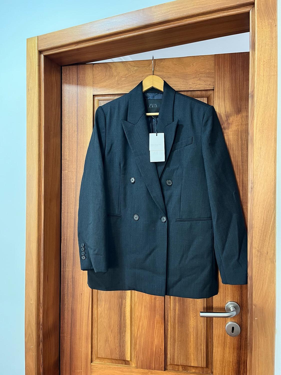 Dark grey double-breasted blazer hanging on a wooden door. Interior label visible with the text ZARA.