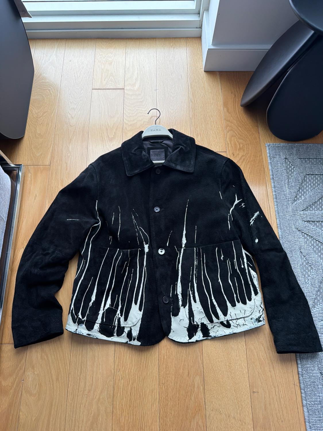 Black jacket with irregular white line design hanging on a Zara hanger over a light wood floor.