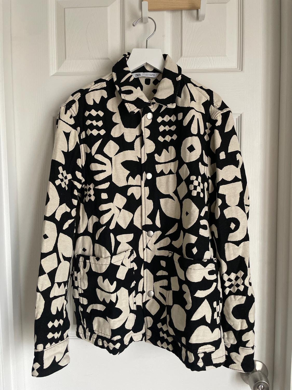 Jacket hanging on a white door, with a black and white abstract print. Visible label with text: "ZARA MAN".