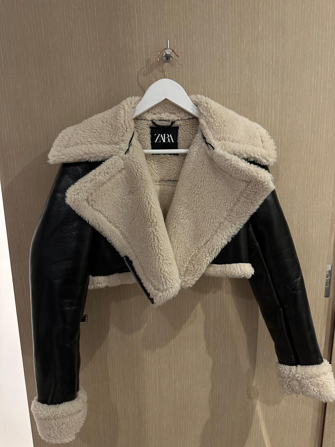 Short black jacket with wide lapels, white fur hem and cuffs, hanging on a white hanger on a wooden door. ZARA label.
