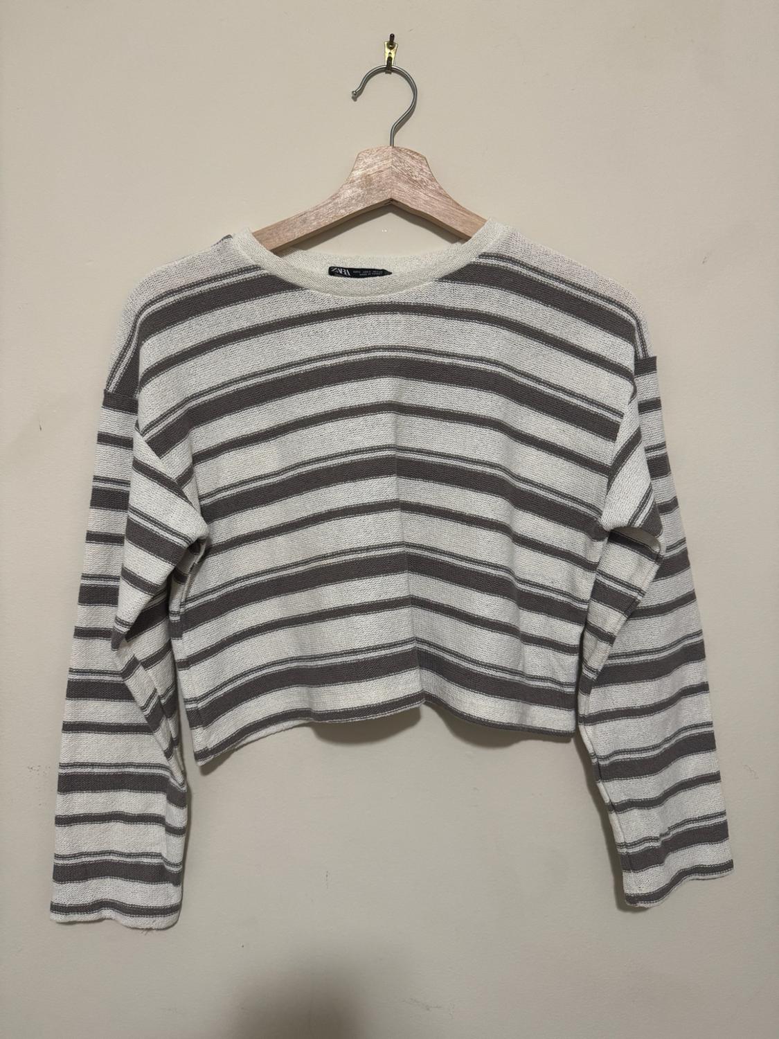 Horizontal striped long-sleeve crop sweater, hanging on a wooden hanger against a plain, light-colored wall. Visible text: ZARA.