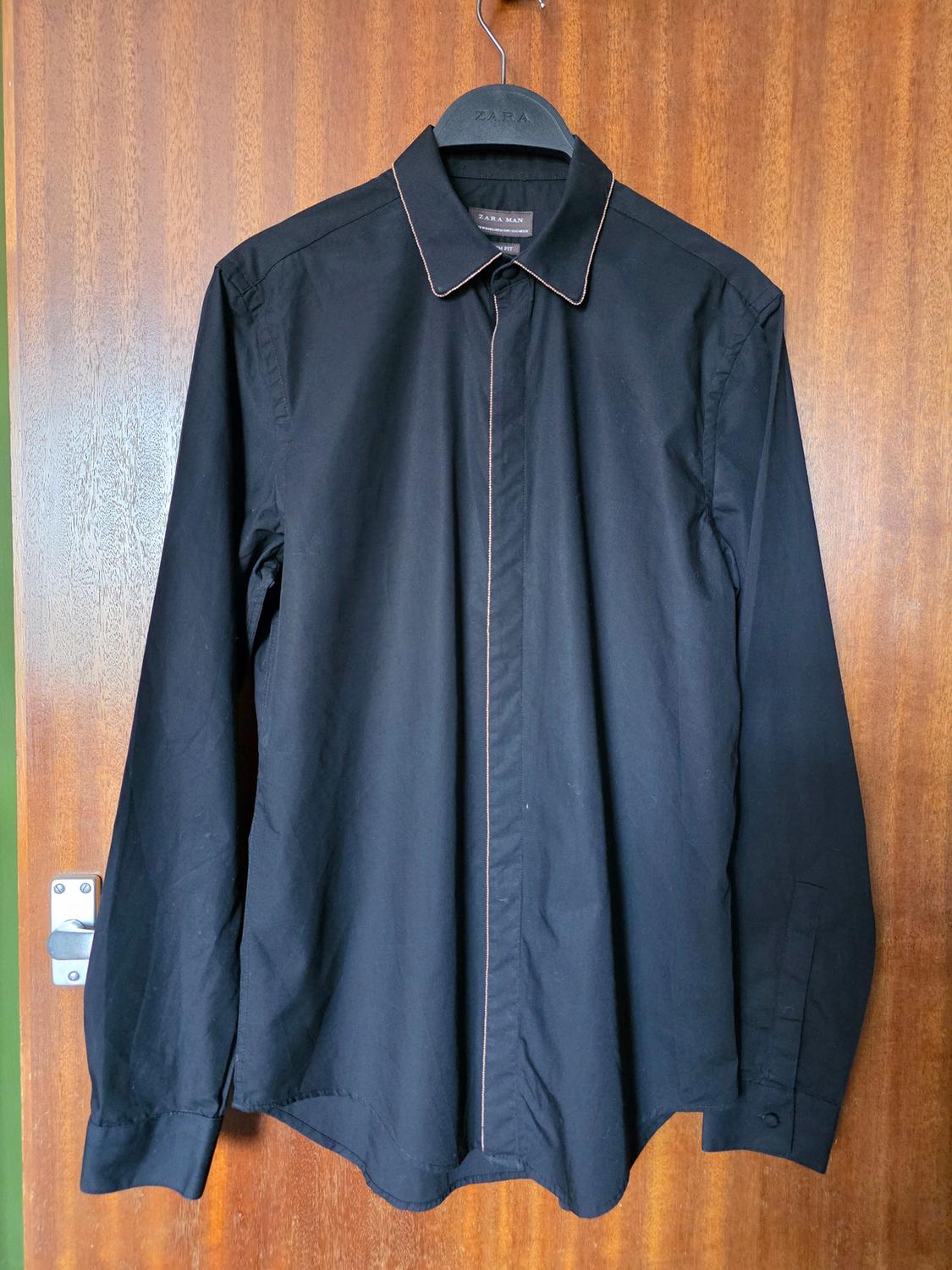 Black long-sleeved shirt hanging on a black hanger with the text "ZARA MAN", on a wooden door.