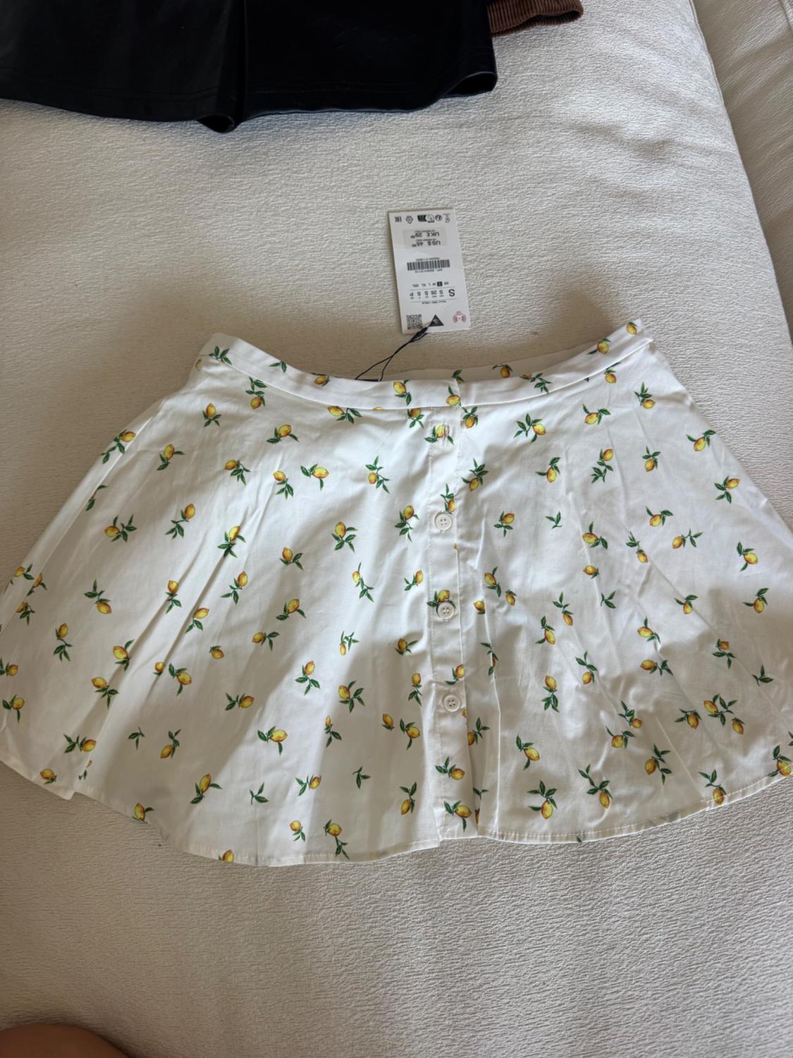 White skirt with a yellow and green lemon print, on a white bed. Visible label with product details.
