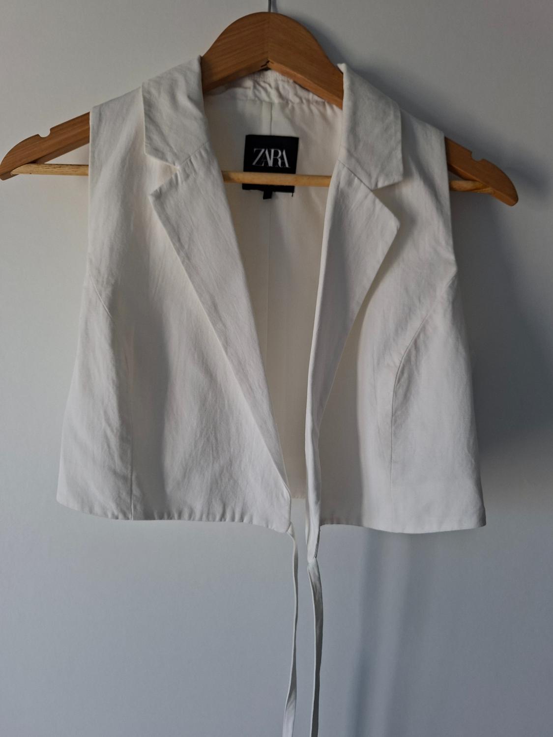White sleeveless waistcoat with a lapel collar and drawstrings at the hem, hanging on a wooden hanger against a white wall. ZARA label.