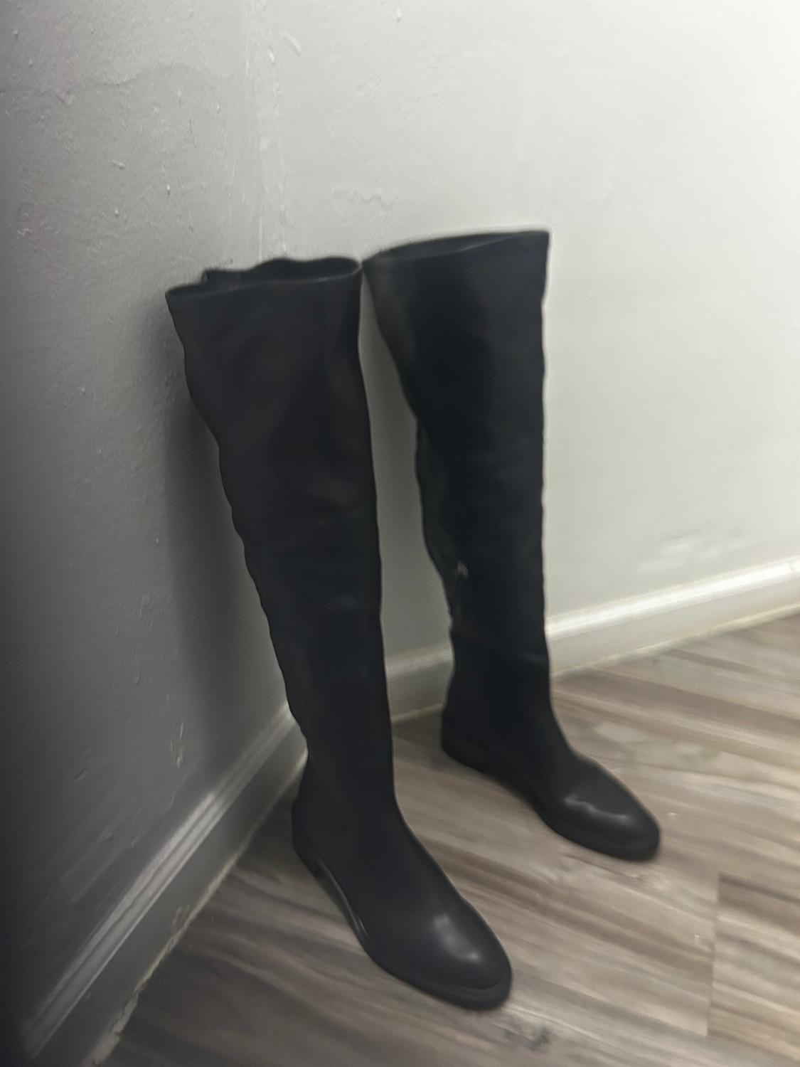Black tall boots leaning in a corner on a light wooden floor, with a white wall background.