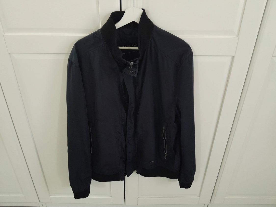 Black jacket hanging on a white hanger in front of a white wardrobe. The jacket has a zip and a high collar.
