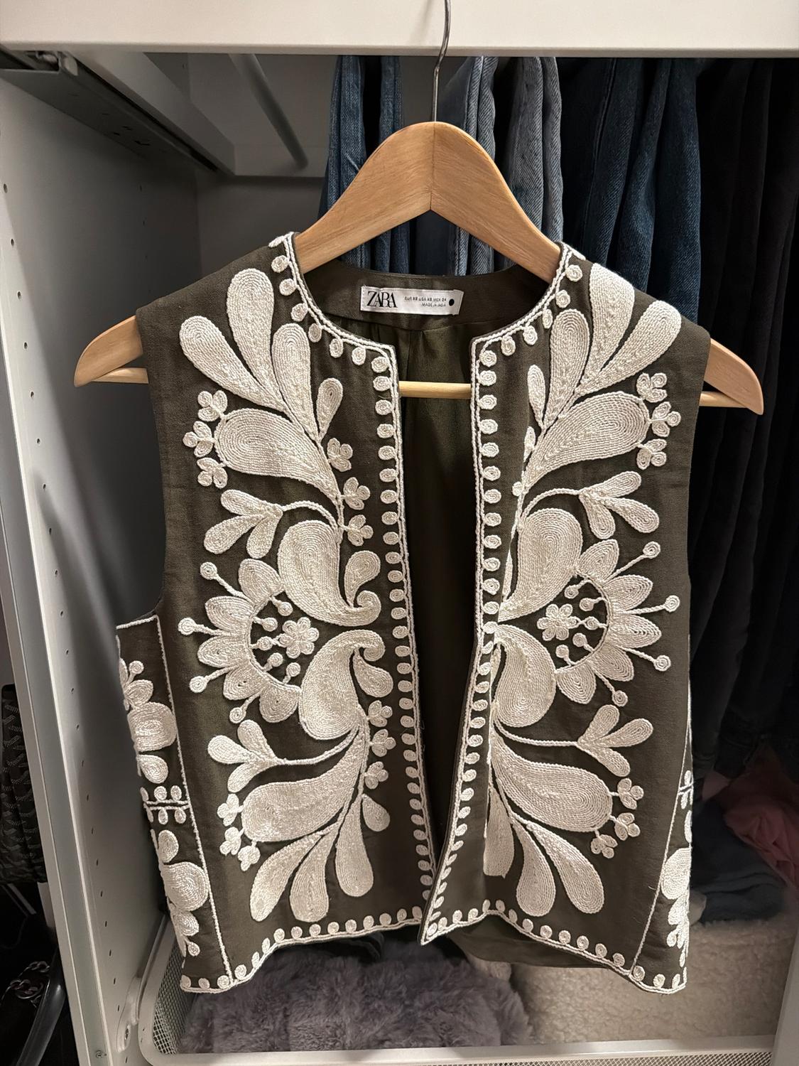 Green waistcoat with white floral embroidery hanging on a wooden hanger inside a wardrobe, Zara label visible.