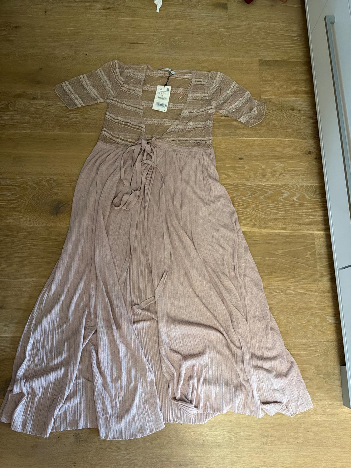 Light pink, short-sleeved maxi dress with a pleated texture, waist tie and visible label, on a wooden floor.