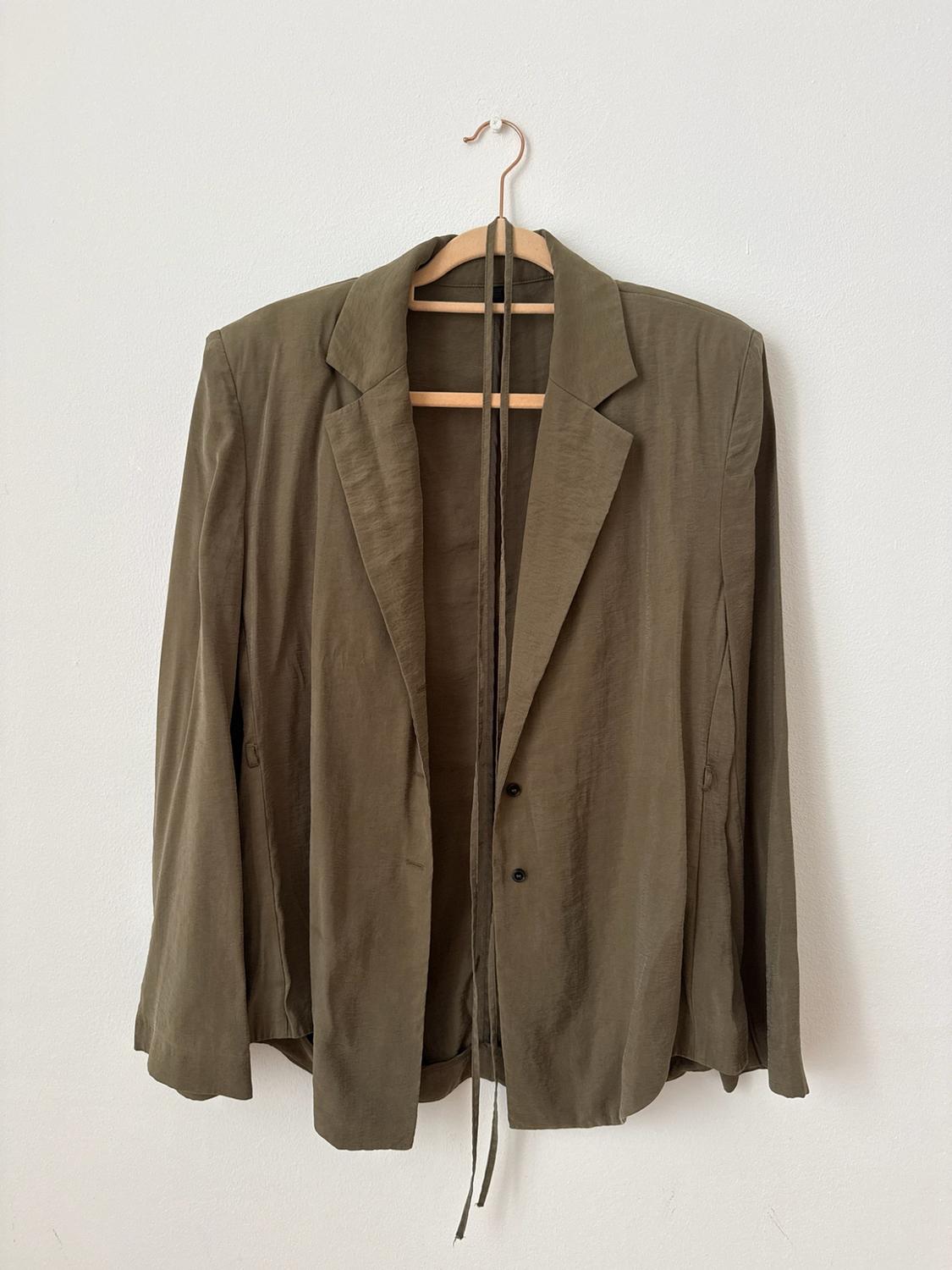 Olive green blazer on a wooden hanger against a white background. It has lapels, buttons, and long ties at the neck.