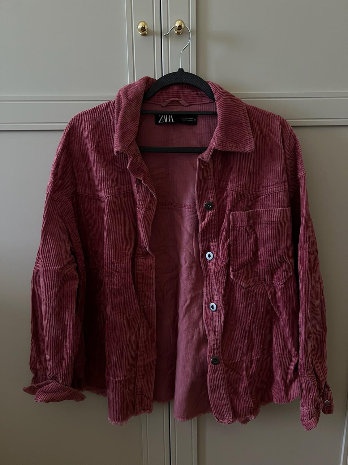 Pink corduroy overshirt with buttons and a frayed hem, hanging on a black hanger in front of a wardrobe. Visible text: ZARA.