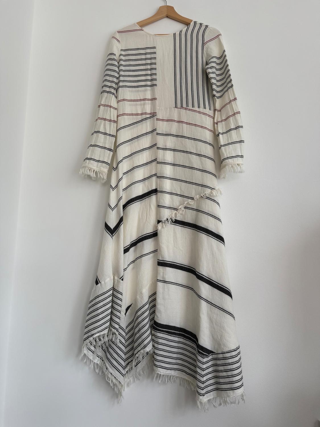 A long dress with black and red stripes on a white background, with fringing on the hem, hanging on a wooden hanger.