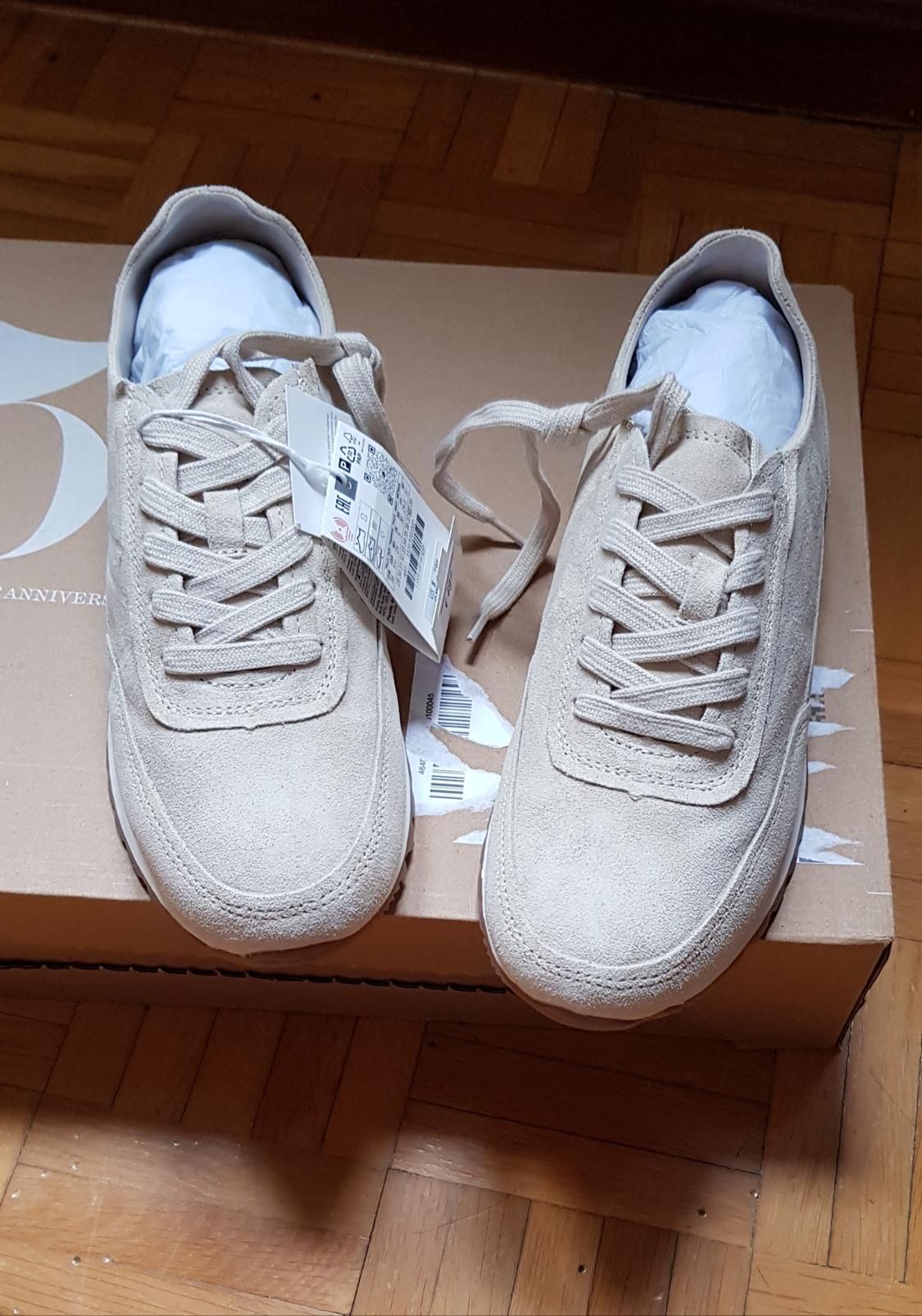 Beige suede trainers with laces on a brown box. Label visible on one trainer. Wooden parquet background.
