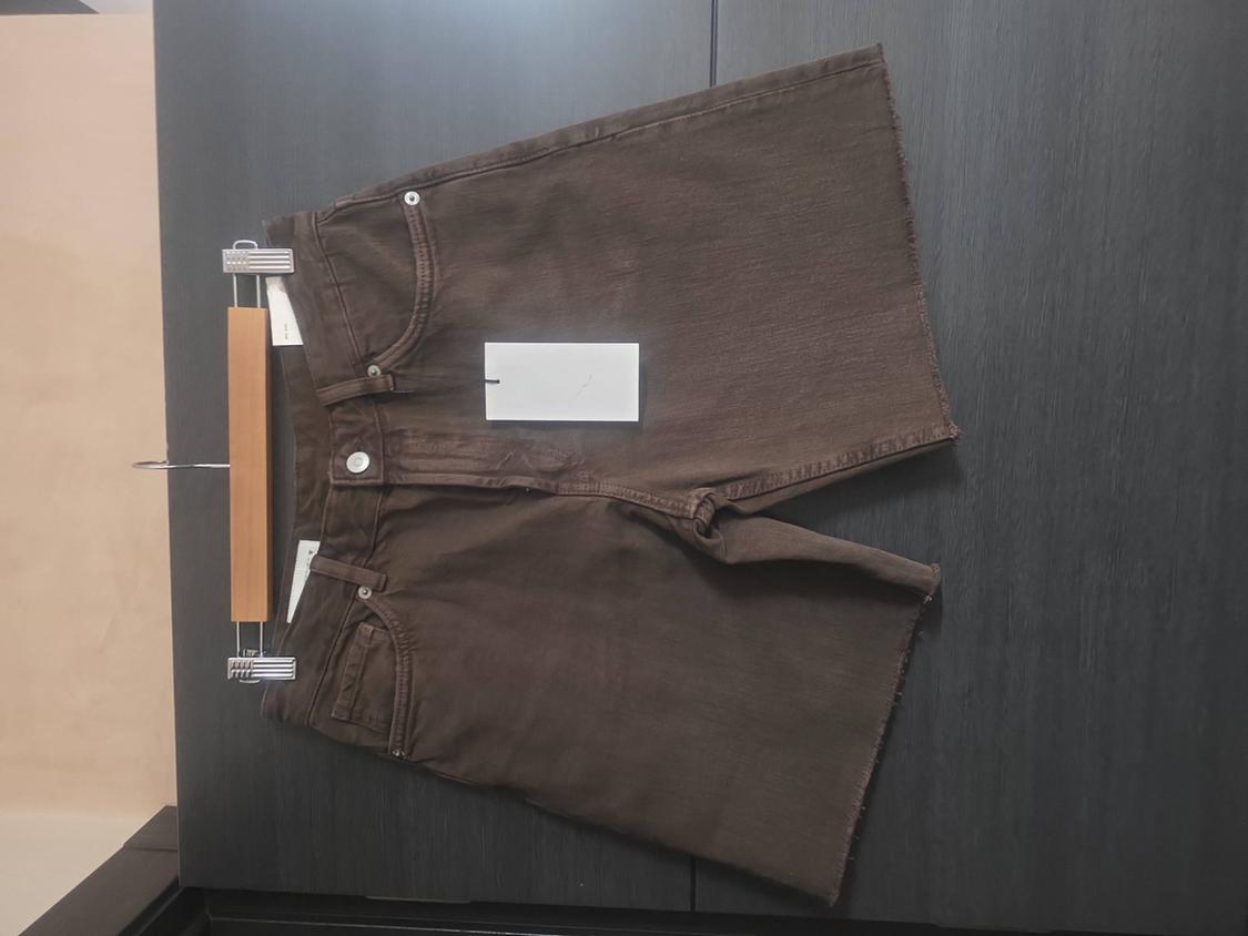 Brown denim shorts with frayed hem and white label, hanging on a wooden hanger against a dark surface.