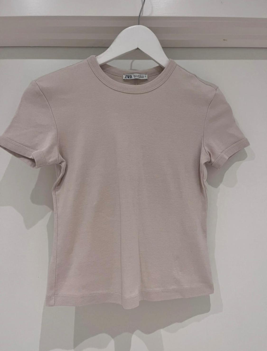Beige short-sleeved T-shirt hanging on a white hanger against a white background. Visible label with text "ZARA".