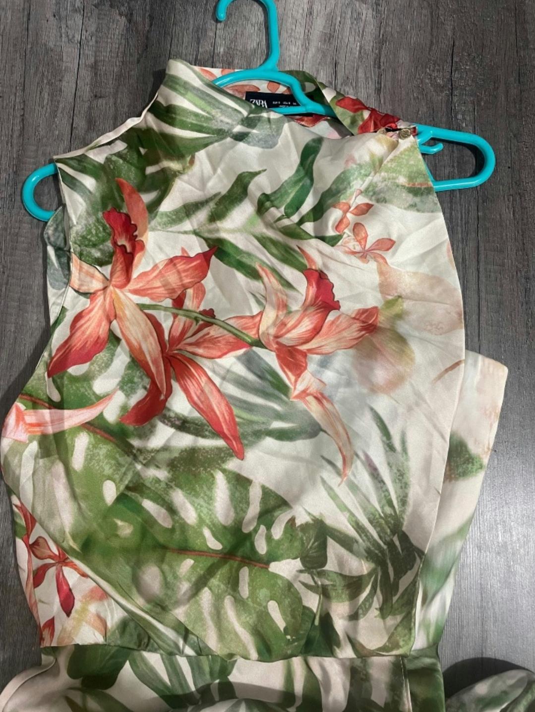 Sleeveless blouse with a tropical floral print in green and red tones, hung on a blue hanger over a grey wooden floor.