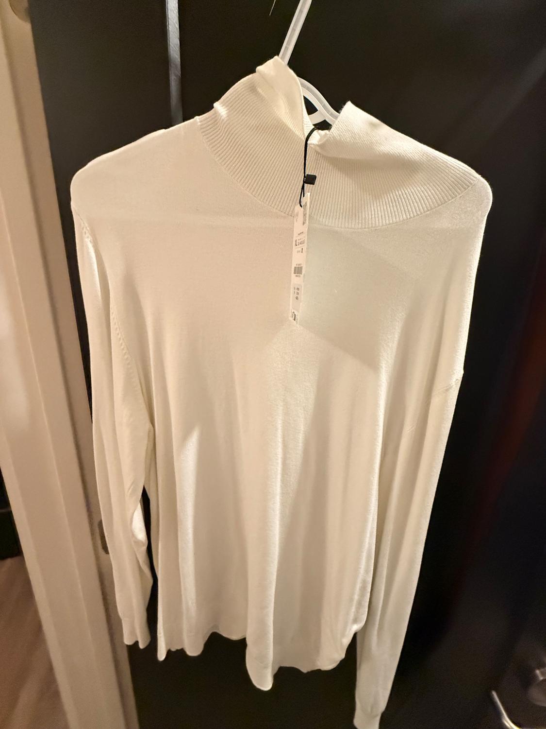 White turtleneck sweater hanging on a hanger, with a visible tag, against a dark background in an indoor setting.