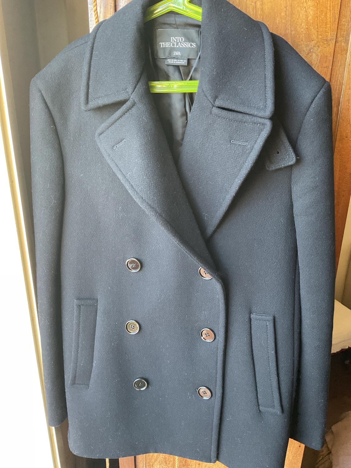 Μαύρο σακάκι με διπλή διαγώνια κούμπωμα σε πράσινη κρεμάστρα, με πέτα και κουμπιά. Εσωτερική ετικέτα με κείμενο "INTO THE CLASSICS ZARA". Φόντο από ξύλο.