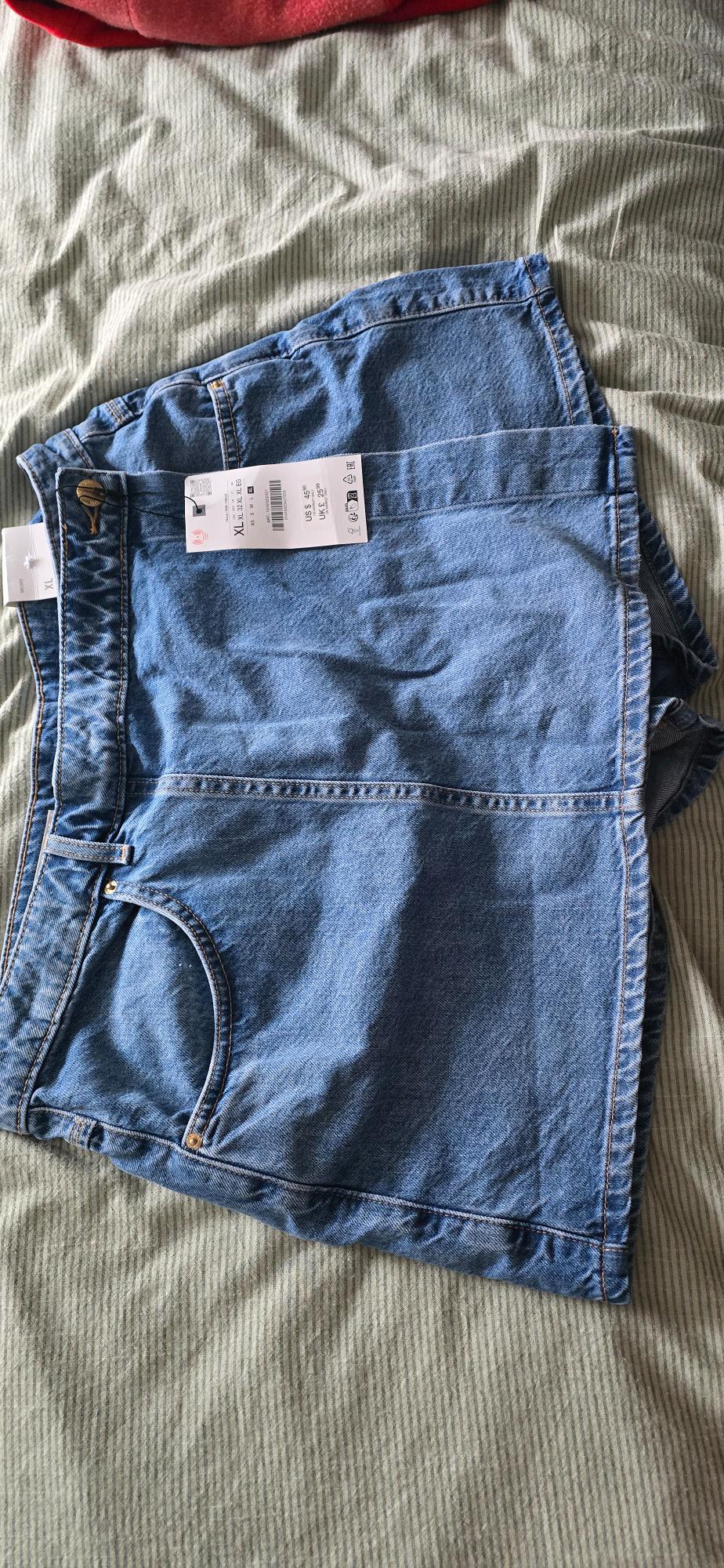Blue denim shorts with price tag and XL size on a bed with a green striped bedspread.