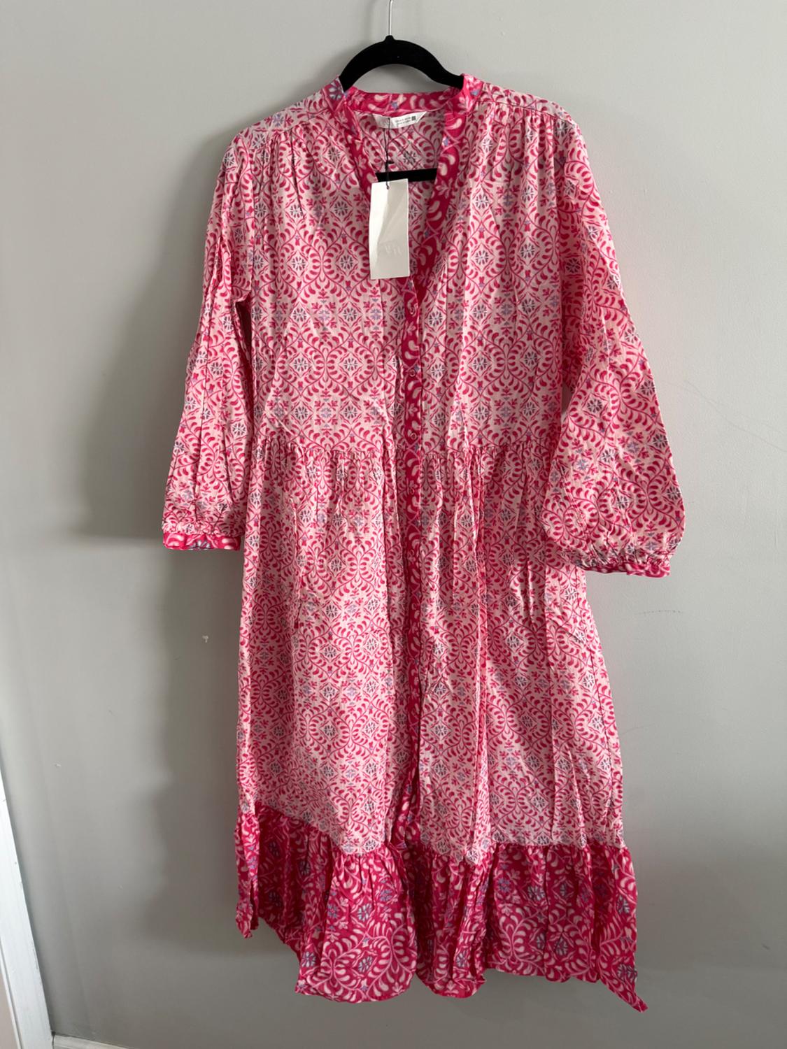 Long dress with floral print in pink and white tones, hanging on a black hanger against a gray wall. Visible tag.