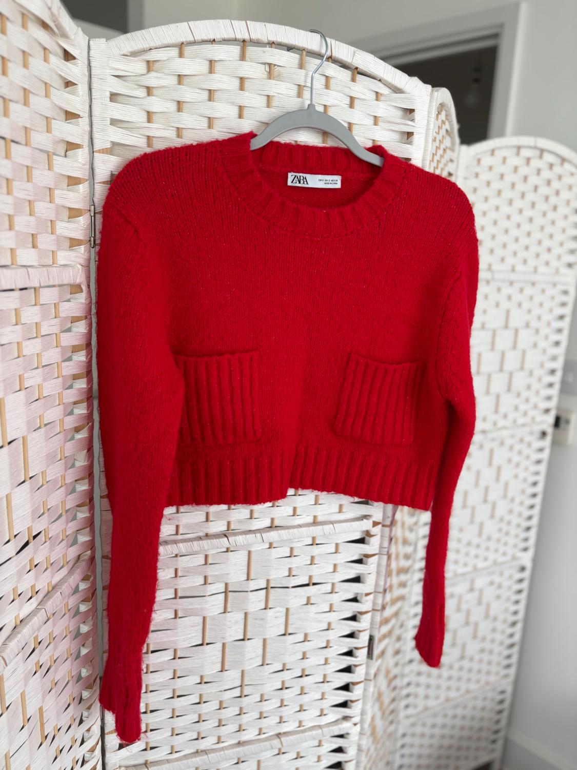 A red jumper with pockets is hanging on a coat stand in front of a white woven screen. The visible label shows the text "Zara".