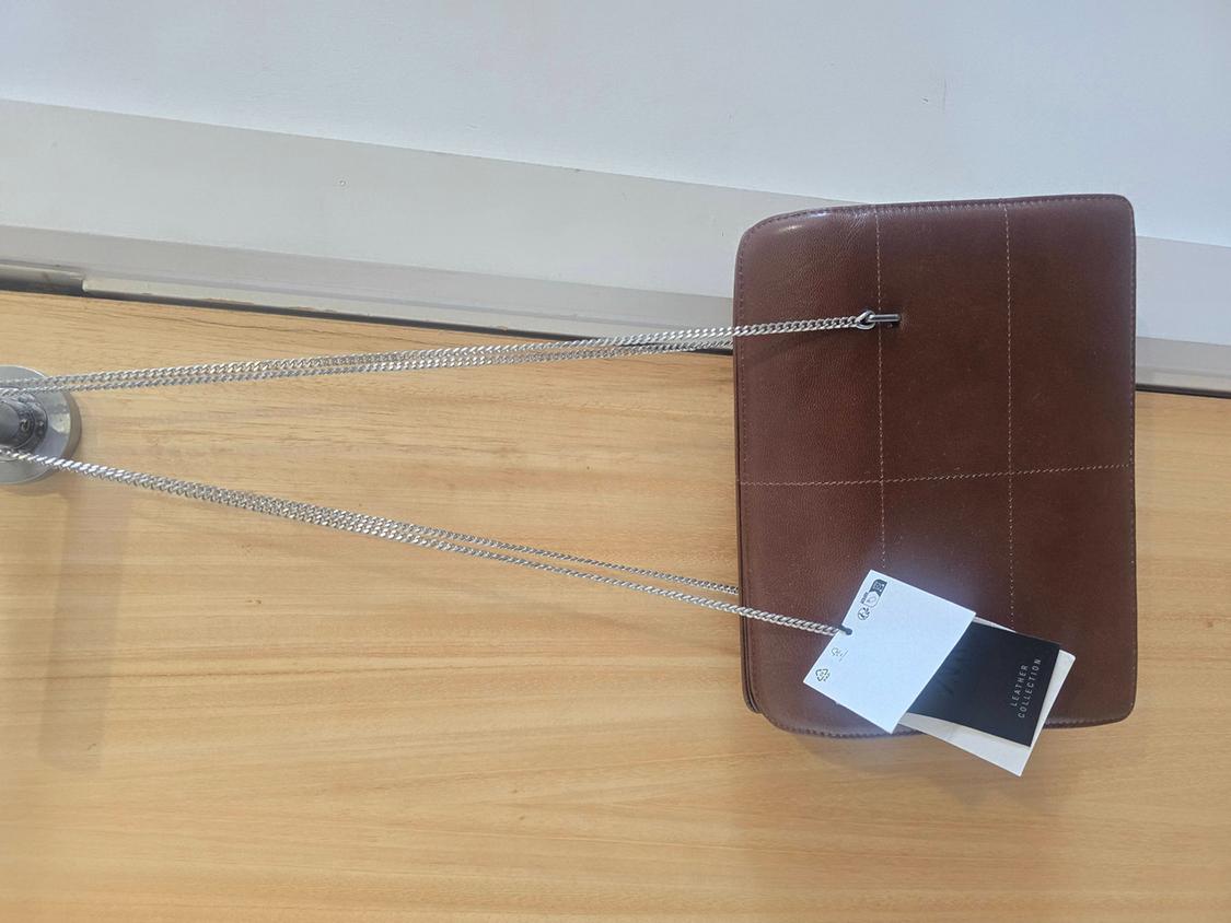 Brown handbag with stitching and a silver chain hanging against a wooden background. Label with the text LEATHER COLLECTION.