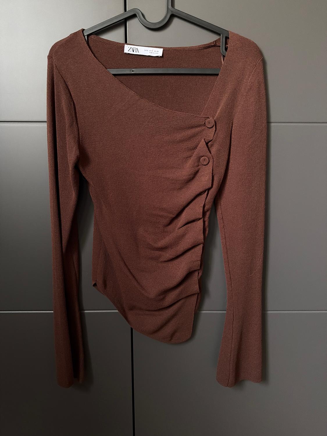 Brown long-sleeved blouse with decorative buttons on the shoulder, hanging on a black hanger against a grey background. Visible tag: "ZARA".