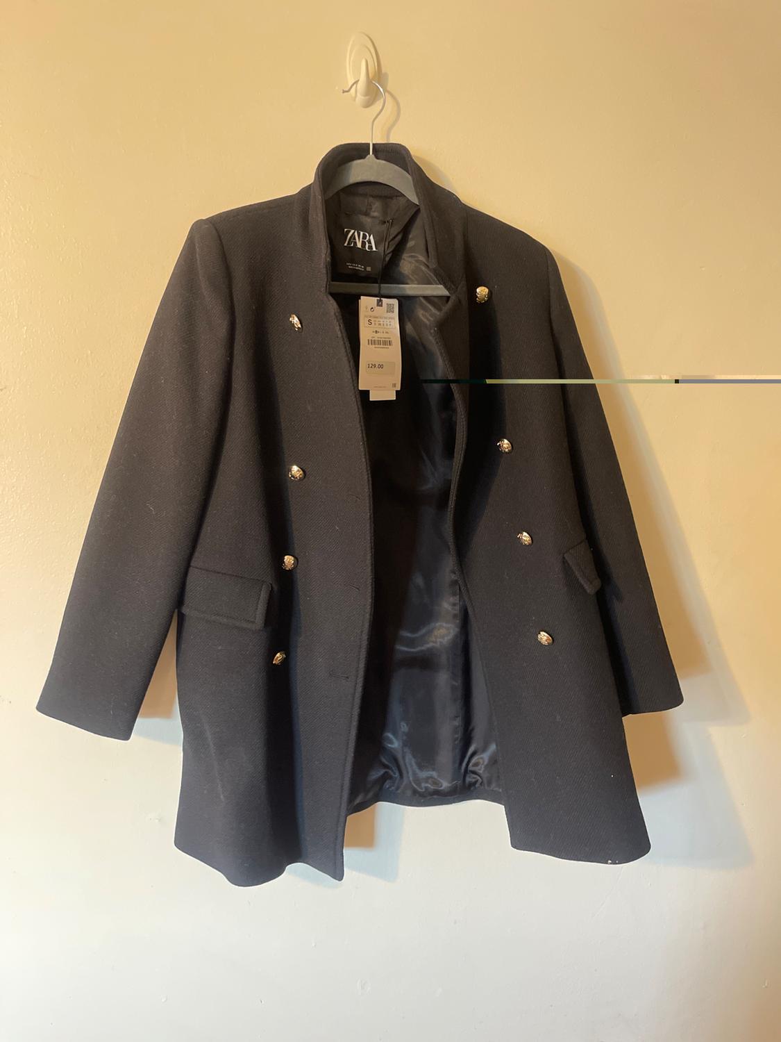 Black coat hanging on a hanger, with gold buttons and a visible tag. White background. Visible brand: ZARA. Price on tag: 129.00.