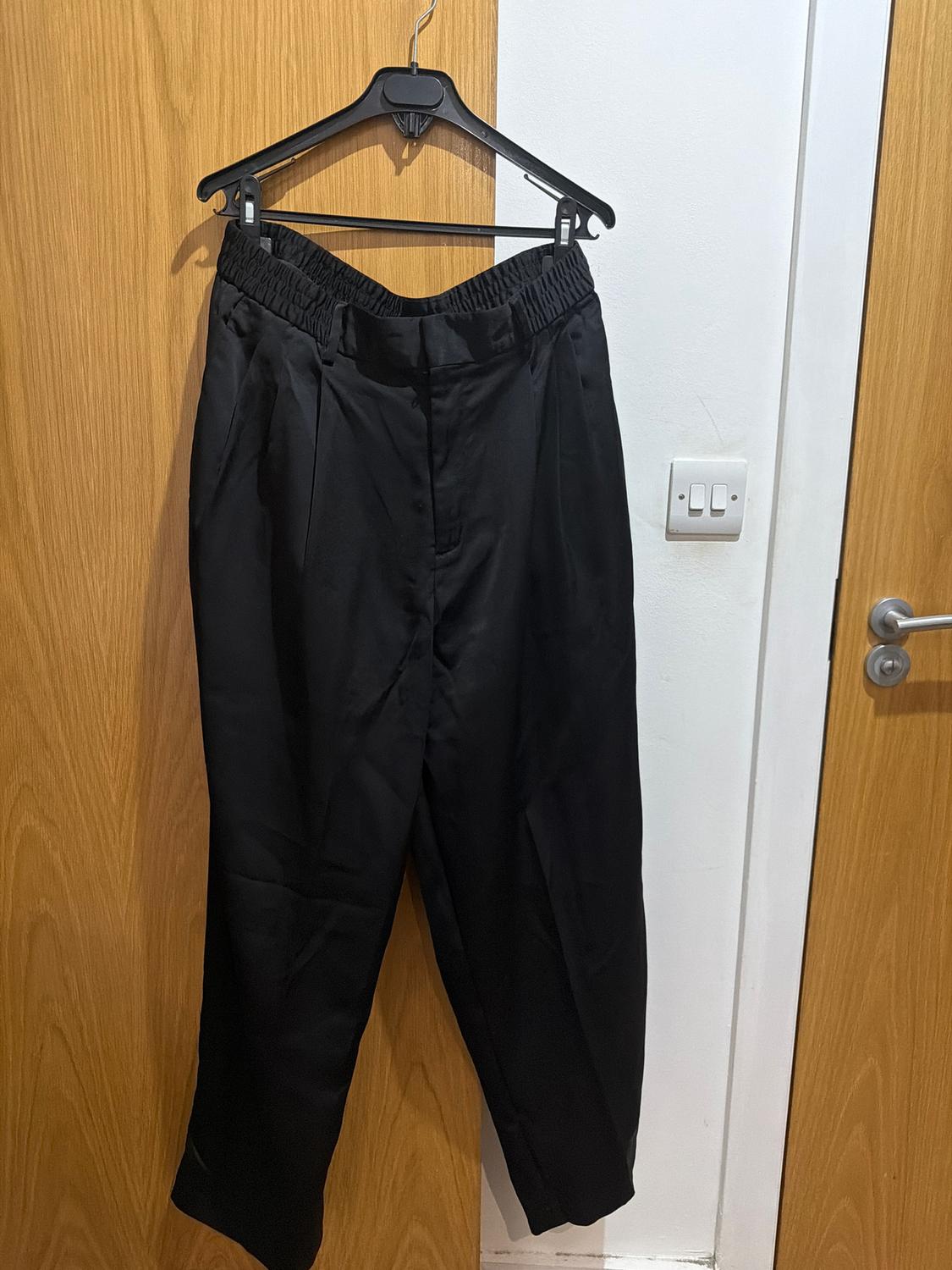 Black trousers hanging on a black hanger over a light wooden door. White background with a visible light switch.