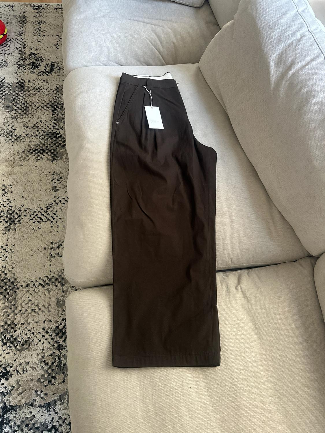 Dark brown pants folded on a comfortable beige sofa. A white label can be seen on the waistband and a rug is on the floor.