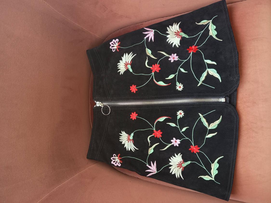 Black mini skirt with floral embroidery and a front zip, photographed from a top-down perspective on a dusty pink armchair.