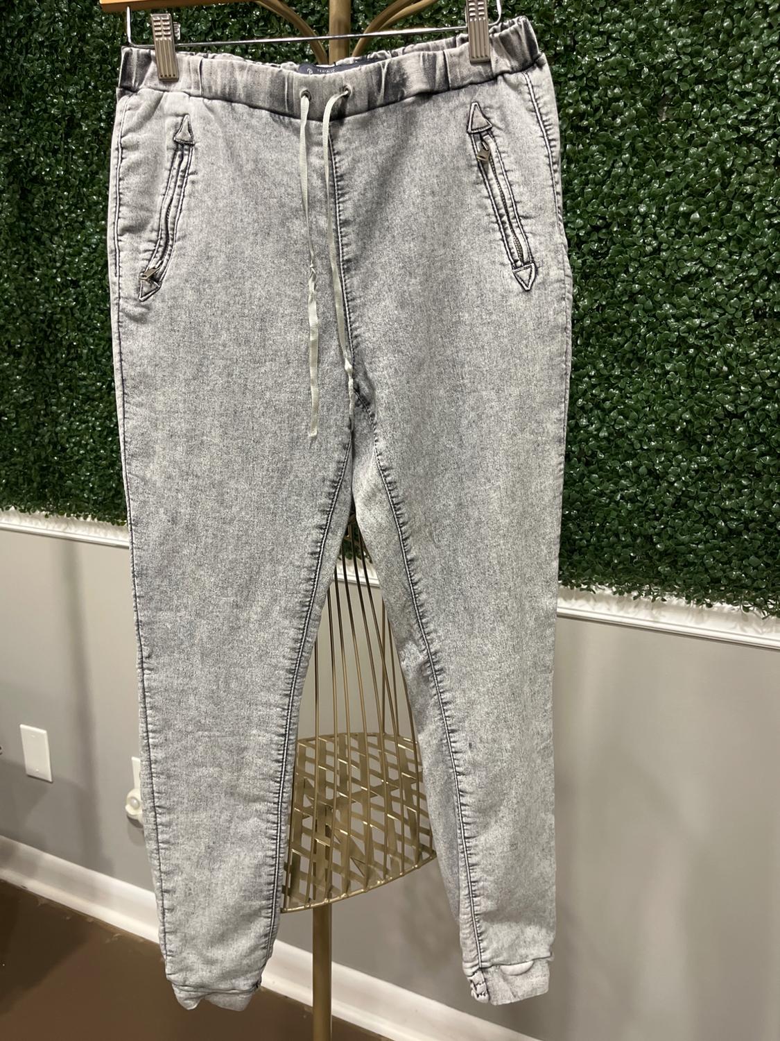 Gray pants hanging on a hanger in front of a green leaf background. They have a drawstring waist and zippered pockets.