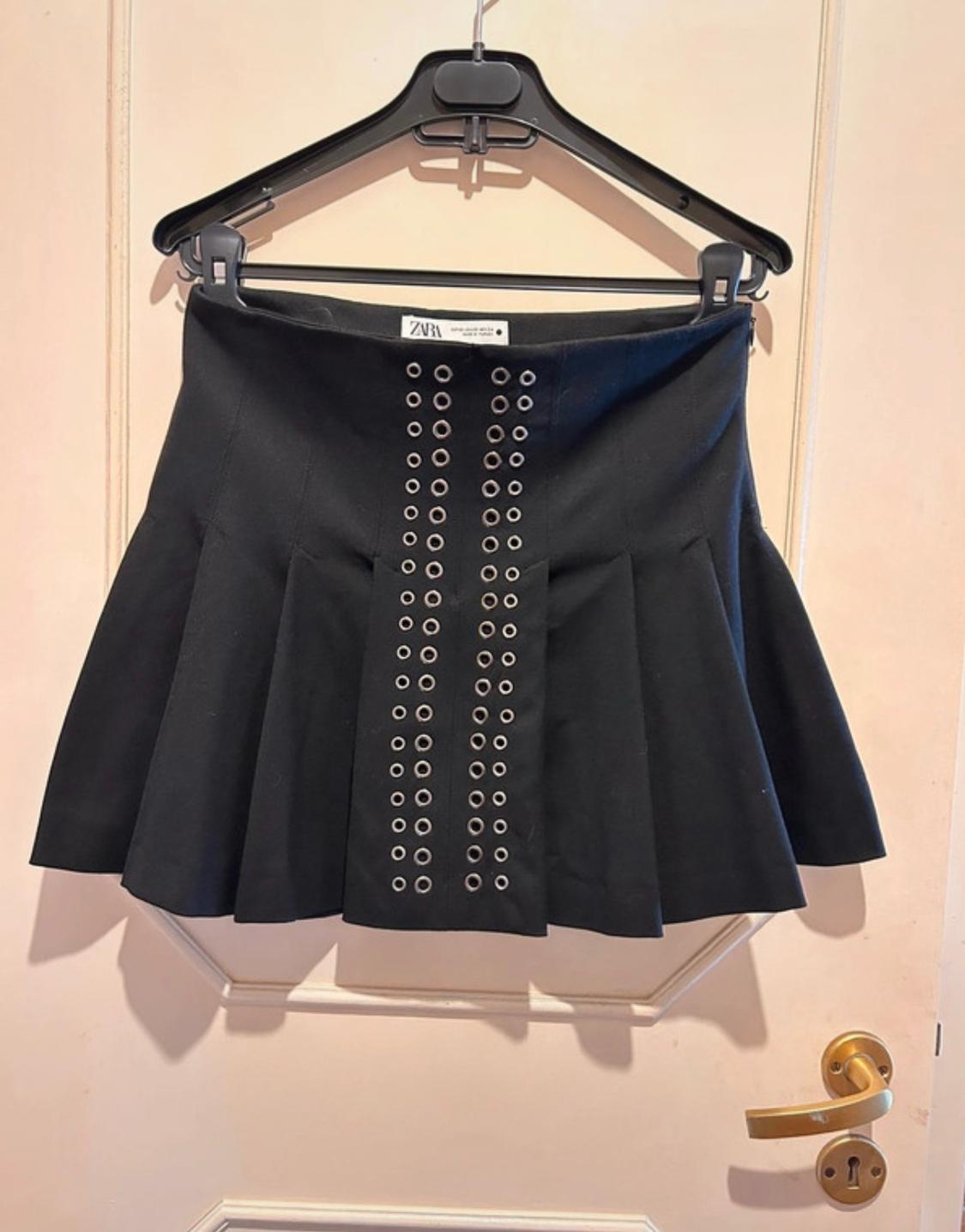 Black skirt with metallic eyelets in the centre, hanging on a black hanger over a white door with a gold handle.