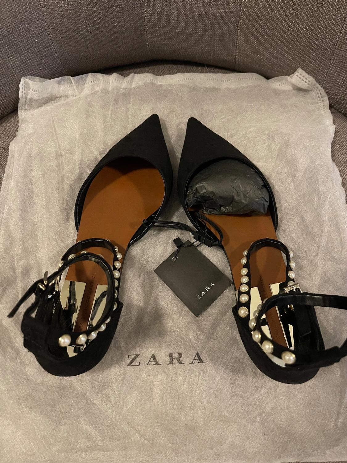 Black pointed shoes with brown interior and pearl straps, on a white bag with the text ZARA and a black label with the text ZARA.