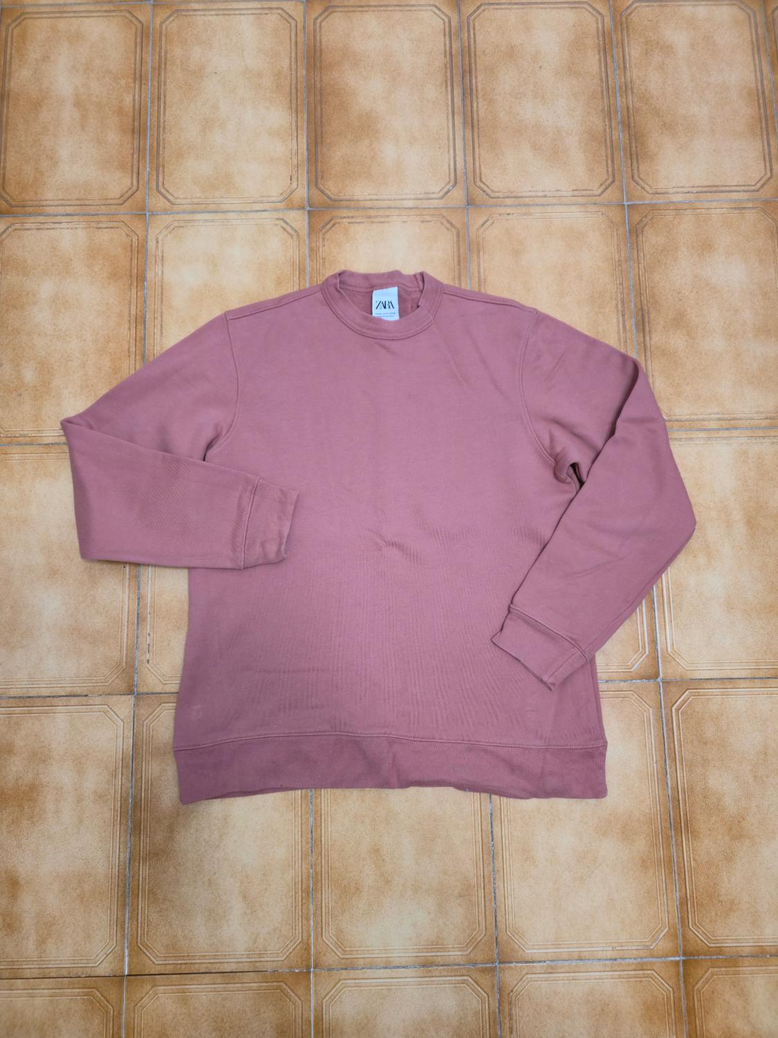 Pink long-sleeved sweatshirt on a brown tiled floor. Visible label with "ZARA" text on the collar.