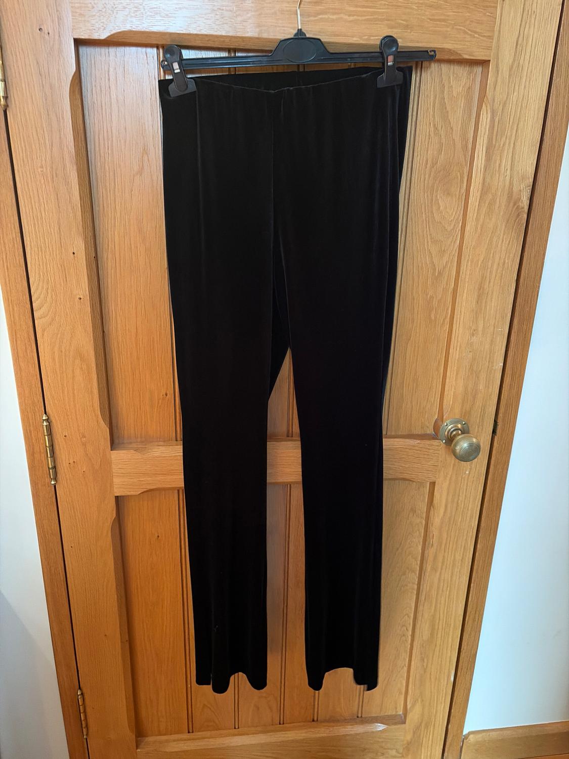 Black velvet trousers hanging on a black hanger over a light wooden door.