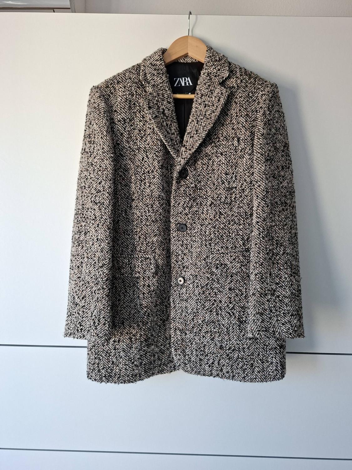 Herringbone-patterned black and white coat hanging on a wooden hanger against a white background. Label with the text ZARA.