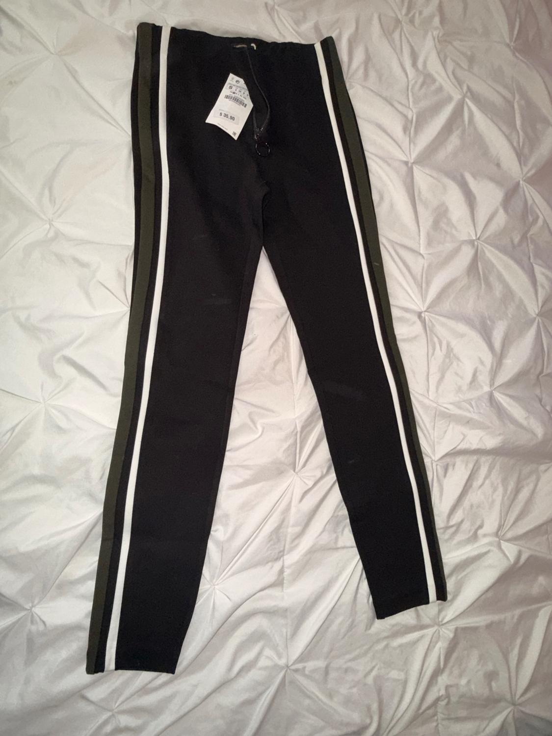 Black pants with white and green stripes on the sides, on a white bedspread. Visible tag with a price of $39.90.