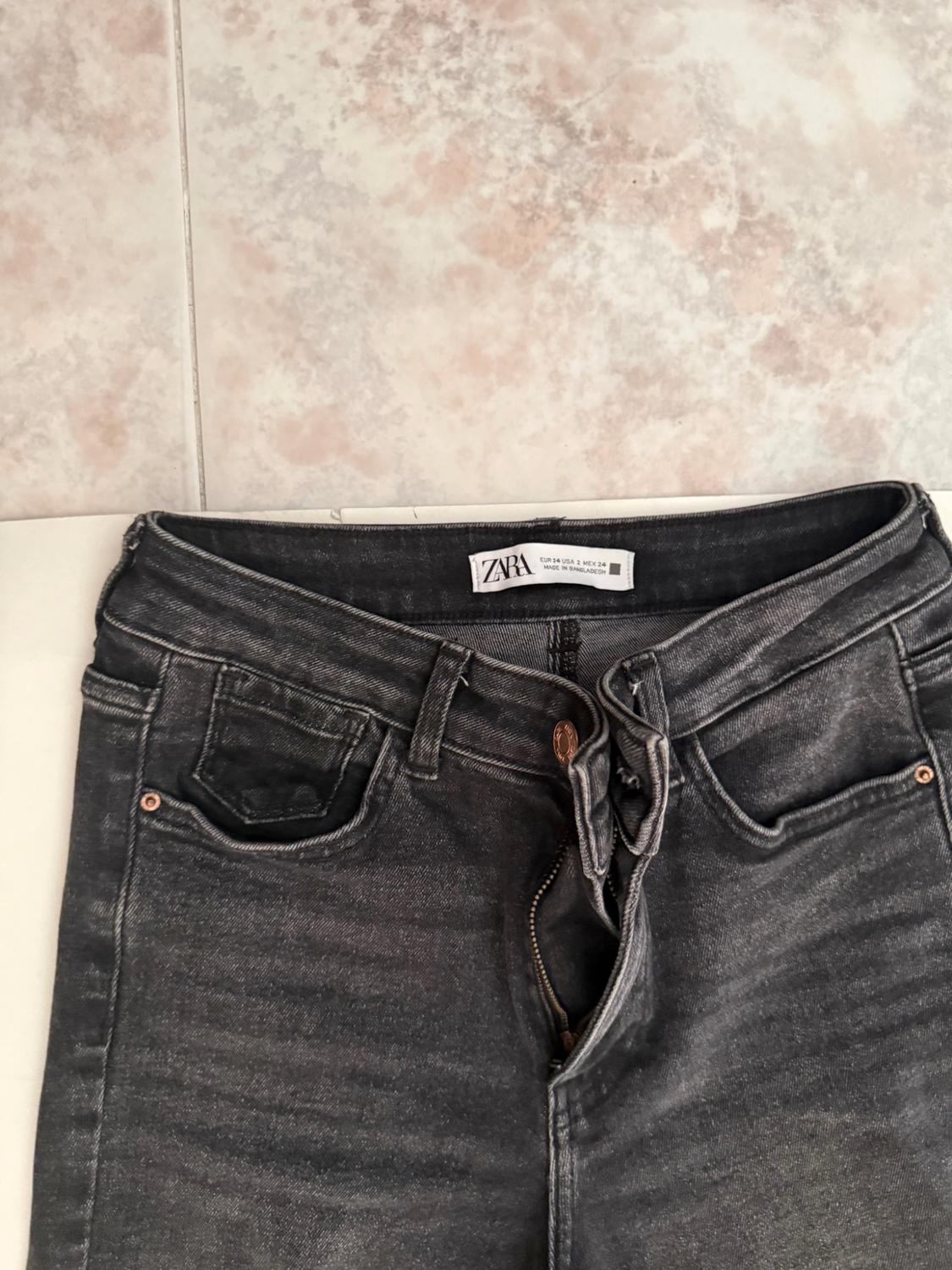 Dark grey unbuttoned jeans on a light background. Visible text: ZARA EUR 34 USA 2 MEX 24 MADE IN BANGLADESH.