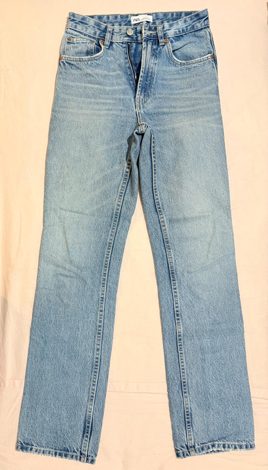 Light blue straight-fit jeans with the ZARA label, unbuttoned and laid out on a wrinkled peach-coloured fabric background.