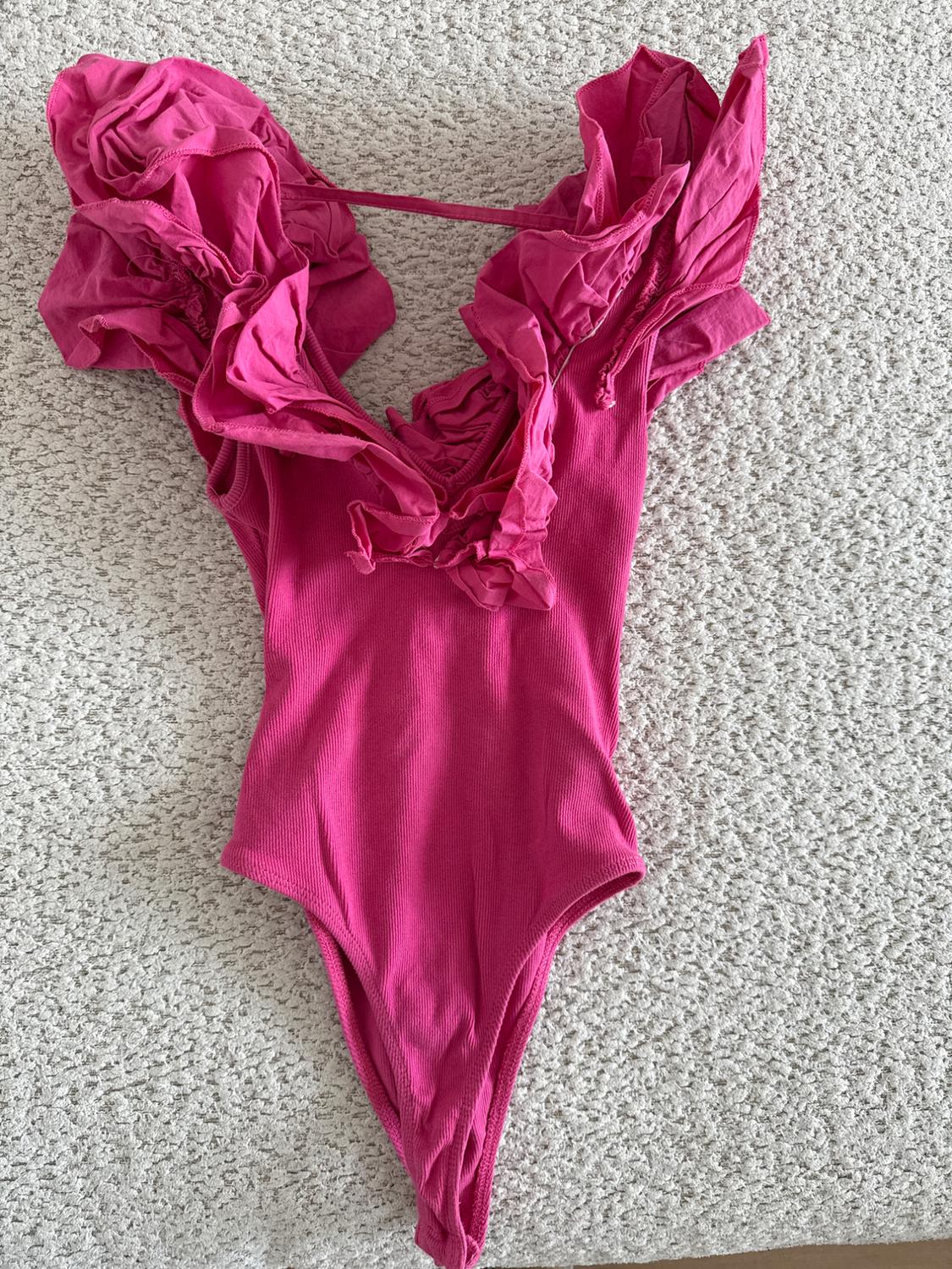 Pink bodysuit with ruffled shoulders, on a light gray textured surface.
