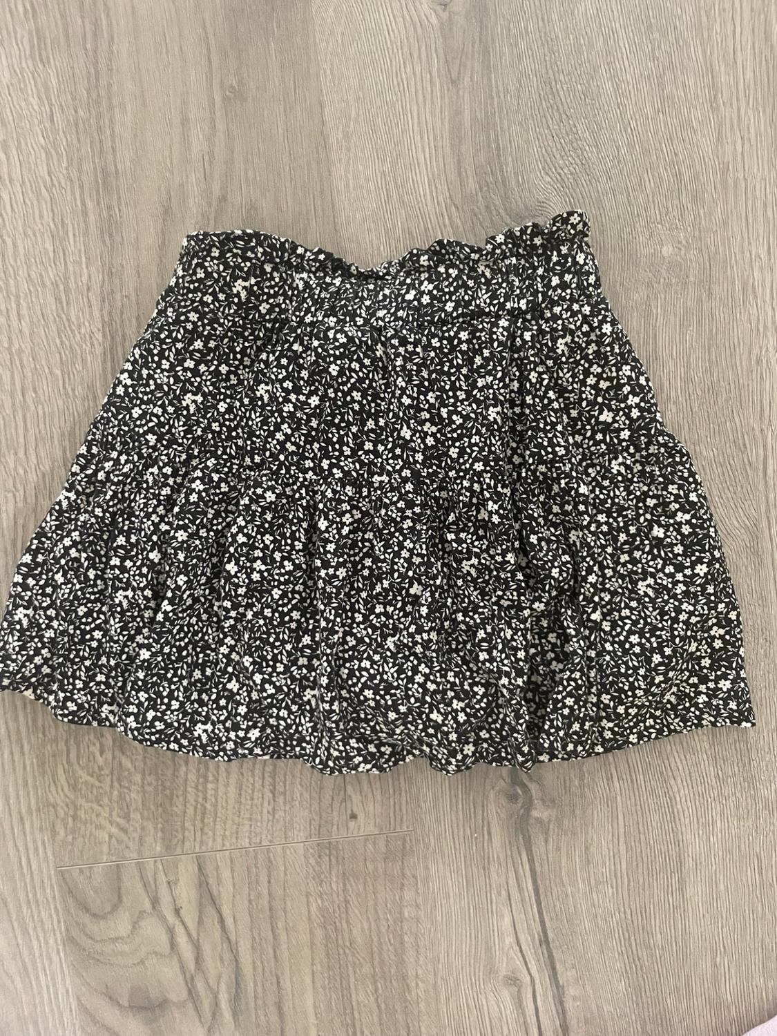 Short black skirt with a white floral print, ruffles, and a gathered waist, on a light wood floor in a casual composition.