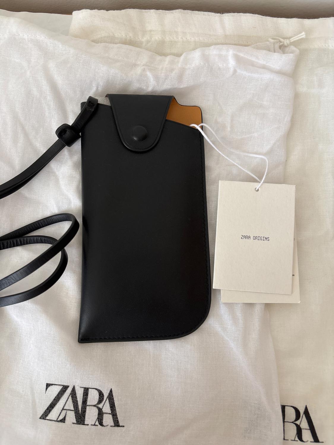 Black case with strap on white fabric with "ZARA" logo. Hanging tag reads "ZARA ORIGINS". Minimalist and elegant atmosphere.