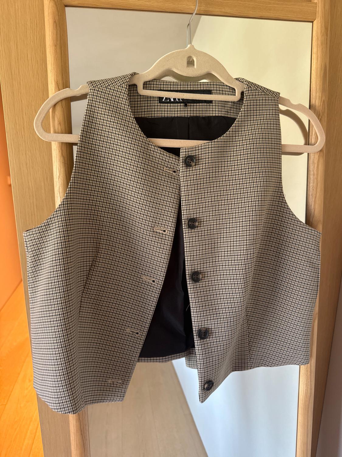 Small black and white checked waistcoat hanging on a wooden hanger, with a mirrored background and a light-coloured wall.