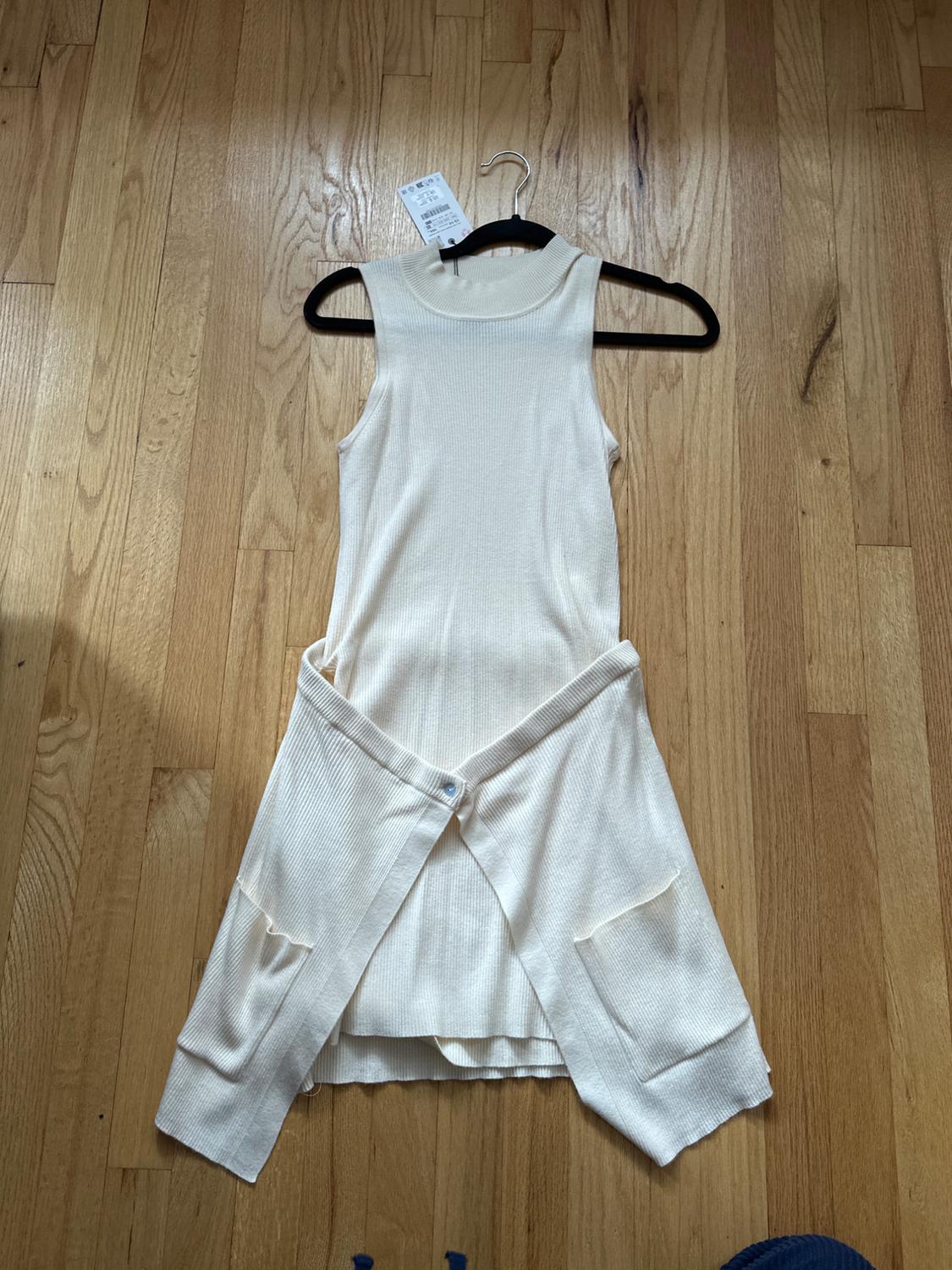 Cream-colored sleeveless dress with a belt, hanging on a black hanger over a light wood floor. Label visible on the hanger.