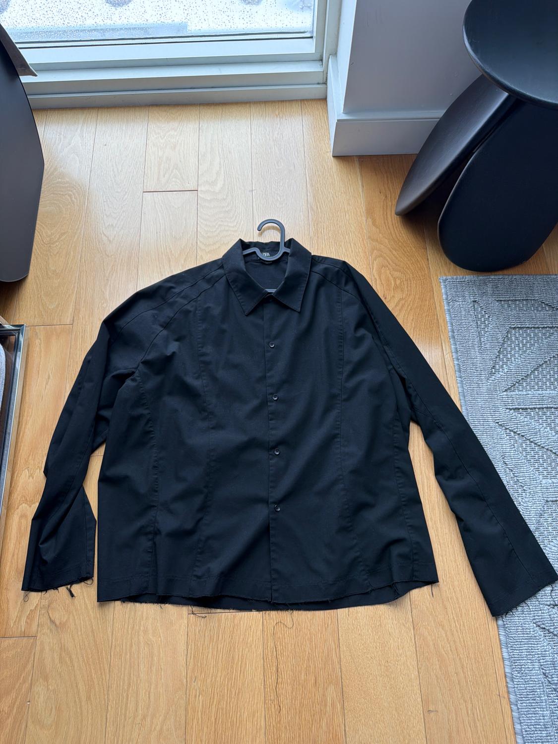 Black long-sleeved shirt on light wood flooring, near a window. The shirt is on a hanger.
