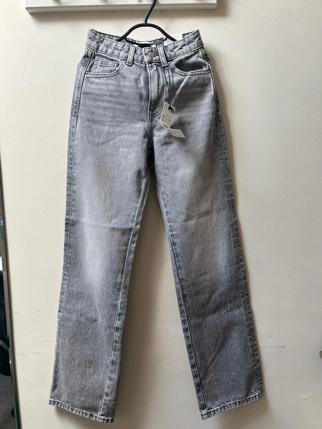 Grey high-waisted, straight-leg jeans with a washed effect, hanging on a black hanger against a light-coloured wall. Casual style.