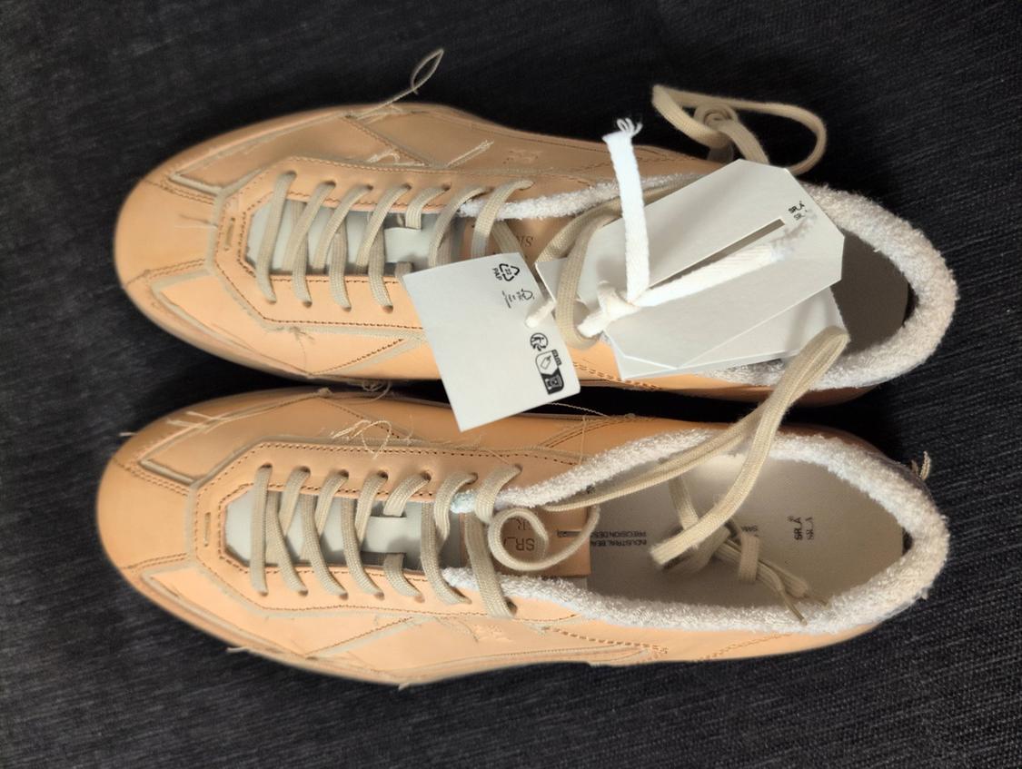 Beige trainers with white laces and swing tags, on a dark background. Interior lined with soft material.