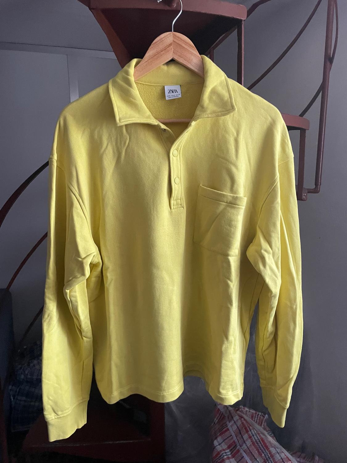 Yellow long-sleeved shirt hanging on a wooden hanger, with a chest pocket and a spiral staircase in the background.