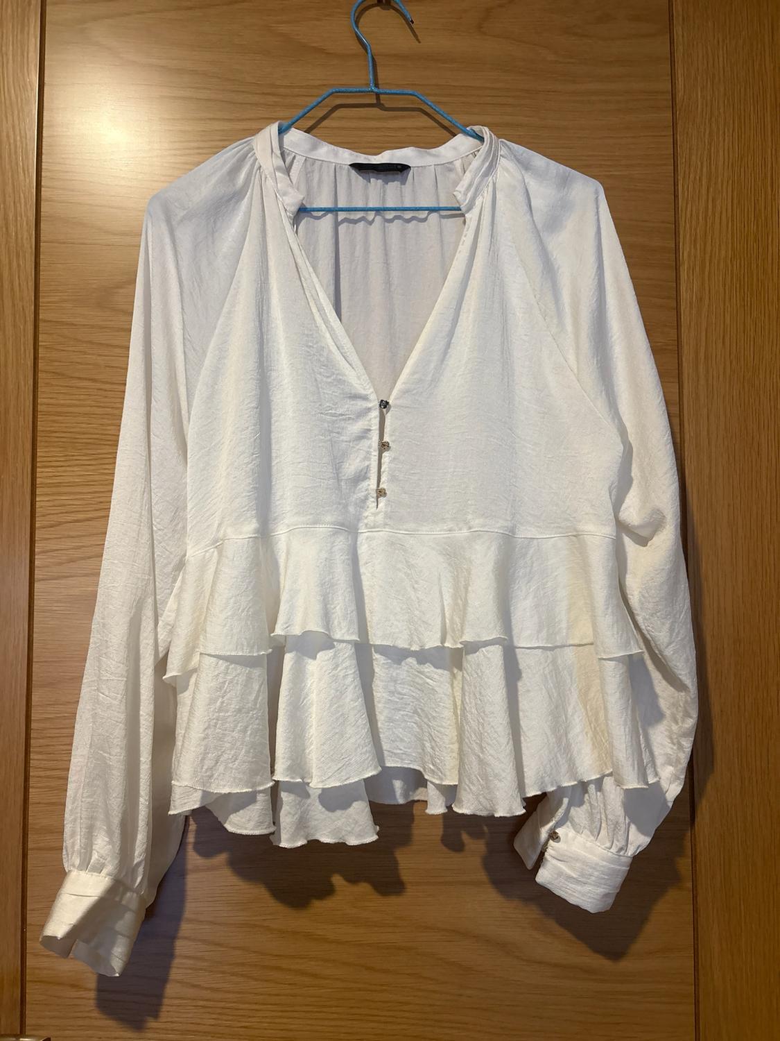 White long-sleeved V-neck blouse with buttons and a frilled hem, hanging on a blue coat hanger in front of a wooden door.