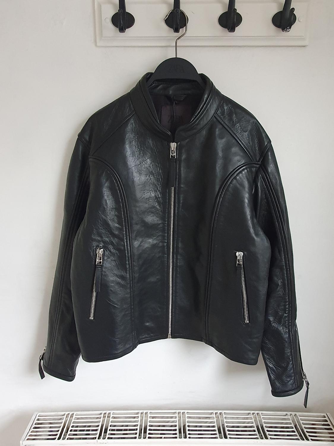 Black leather jacket hanging on a hanger, with silver zips on the pockets and sleeves, against a white background.